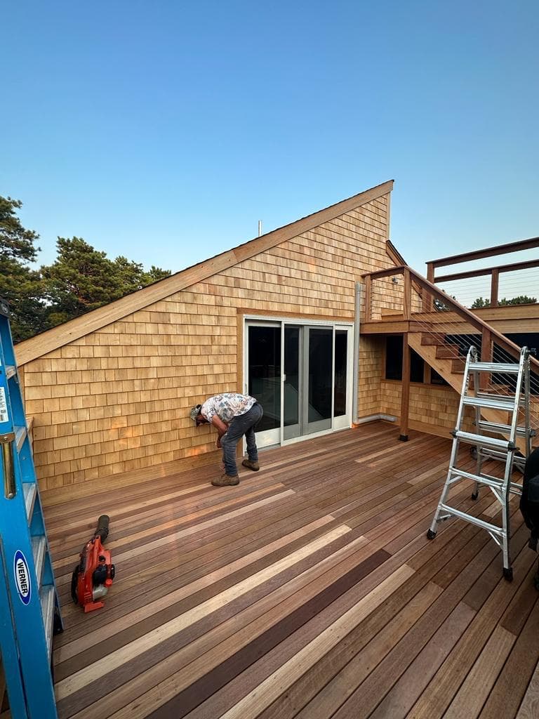 An AKL carpenter kneeling to fasten decking on a mahogany porch beside a cedar-shingle wall