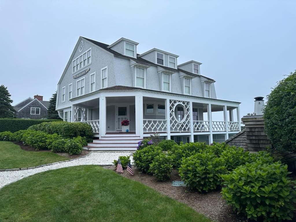 Grey cedar-shingle Cape Cod residence with a wraparound farmer's porch, white trim and clipped hedges